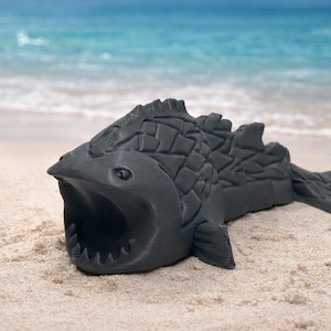 May include: A dark gray, stylized fish sculpture with an open mouth, resting on a sandy beach with the ocean in the background. The fish has textured scales and fins, and is likely a decorative item.