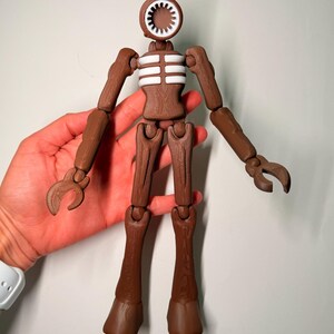 May include: A brown articulated figure with a siren head and claw-like hands. The figure has a white and brown torso and is held in a person's hand. The figure's head is a speaker-like shape with a white interior.