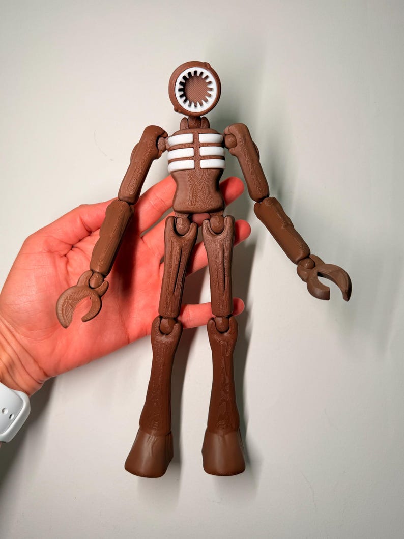 May include: A brown articulated figure with a siren head and claw-like hands. The figure has a white and brown torso and is held in a person's hand. The figure's head is a speaker-like shape with a white interior.