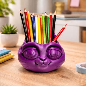 May include: A purple, cat-shaped pencil holder filled with a variety of colored pencils. The holder has large, round eyes and a smiling face. The pencils are arranged vertically, with the tips visible.