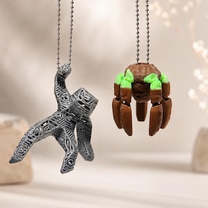 May include: Two pendants on silver-toned ball chains. One is a gray, textured, geometric figure with an outstretched arm. The other is a brown, rounded, multi-legged creature with bright green accents on its upper joints and a textured surface.