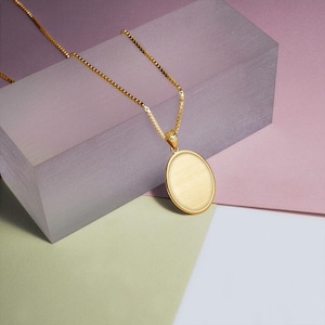 Peut inclure: Un collier doré avec un pendentif ovale. Le pendentif et la chaîne sont en métal brillant. Le collier est présenté sur un fond multicolore avec des nuances de rose, de violet, de vert et de blanc.