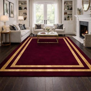May include: A rectangular burgundy rug with a gold border in a living room setting. The rug is the focal point, with a gold border that frames the burgundy center. The room features white furniture, a fireplace, and a chandelier.