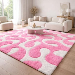 Può includere: Un grande tappeto rettangolare con un vivace motivo astratto rosa e bianco. Il design del tappeto presenta forme organiche e fluide. La stanza comprende anche un divano bianco, una poltrona e un tavolino, creando uno spazio moderno e invitante.