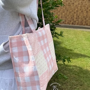 May include: A pink and white gingham tote bag with patchwork details. The bag features a variety of patterns, including gingham, polka dots, and a wave-like design. The bag has long straps and a rectangular shape. The text "THREADED BY EM" is visible on the bag.