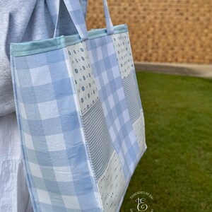 May include: A tote bag featuring a patchwork design of light blue and white fabrics. The pattern includes a large gingham check, a thin grey and white stripe, and a white fabric with small blue floral motifs. It has two matching fabric straps.