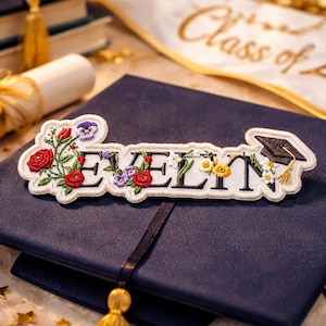 Può includere: Toppa ricamata con il nome "EVELYN" in lettere nere, ornata da rose rosse, fiori gialli e un cappello di laurea. La toppa è posta su un cappello di laurea blu navy, con un nappa dorata e un diploma arrotolato sullo sfondo.
