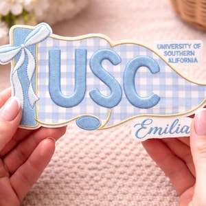Könnte beinhalten: Ein hellblau-weiß besticktes Wimpel mit den Buchstaben "USC" in Hellblau. Der Wimpel hat ein Vichy-Muster, eine Schleife und die Wörter "University of Southern California" und "Emilia".