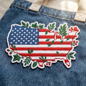 May include: Embroidered patch in the shape of the United States, featuring the American flag design. The patch is outlined with a white border and adorned with green leaves and red berries, creating a patriotic and natural aesthetic. The patch is placed on a blue denim background.