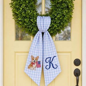 May include: A green boxwood wreath adorned with a light blue and white gingham fabric sash. The sash features an embroidered corgi holding a small American flag on one side and a large, dark blue embroidered letter K on the other side.