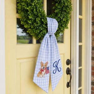 May include: A green boxwood wreath adorned with a light blue and white gingham fabric sash. The sash features an embroidered corgi holding an American flag and a large navy blue script letter K, hanging vertically from the center of the wreath.