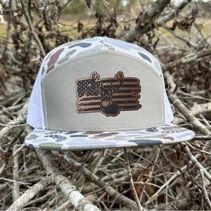 May include: A camouflage-patterned trucker hat with a tan front panel. The front features a brown leather patch with a deer silhouette superimposed over a distressed American flag design. The hat has a white mesh back.
