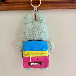 Puede incluir: Un llavero de peluche con un conejito gris claro y una mochila colorida. La mochila tiene una parte superior azul, una cremallera amarilla y una parte inferior rosa. El llavero tiene un anillo dorado y una etiqueta que dice "POP MART".