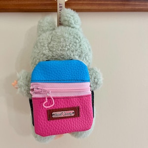 Puede incluir: Un llavero de oso de peluche verde claro con una pequeña mochila colorida. La mochila es azul y rosa con una cremallera rosa y una pequeña etiqueta rectangular "Handmade". El llavero está sujeto a un anillo dorado.