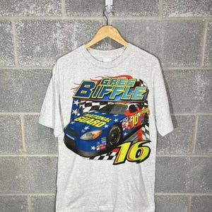 May include: A grey t-shirt displaying a race car graphic with the text "GREG BIFFLE" and the number "16". The car is blue with red and yellow details. The t-shirt is suspended on a wooden hanger against a brick wall.