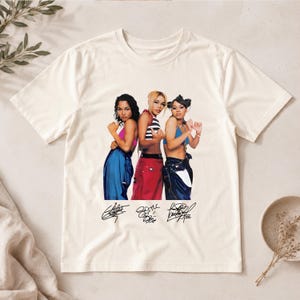 May include: A cream-coloured short-sleeve t-shirt featuring a graphic print of three individuals posing in colourful tops and baggy trousers. Below the figures are three distinct black handwritten-style signatures printed on the fabric.