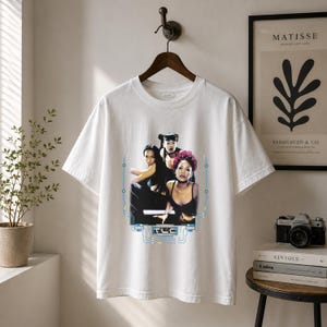 May include: A white short-sleeve t-shirt featuring a graphic print of the music group TLC. The design includes a photograph of the three members, the text TLC in a stylized blue font, and surrounding blue circuit-like line graphics.