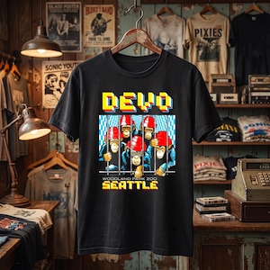 May include: A black t-shirt featuring a graphic of five chimpanzees wearing red hats behind blue and white bars. The text DEVO appears in yellow above the graphic, and the text WOODLAND PARK ZOO SEATTLE appears in yellow below the graphic.