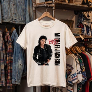 May include: A cream-colored short-sleeve t-shirt featuring a graphic of Michael Jackson wearing a black jacket. The text "BAD" is printed in red above the image, and "MICHAEL JACKSON" is printed vertically in black capital letters to the right.