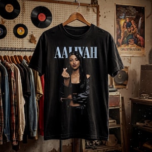 May include: A black short-sleeve t-shirt featuring a graphic print of Aaliyah making a finger heart gesture. The name AALIYAH is printed in large, light blue capital letters above the image. The shirt has a crew neckline and a standard fit.