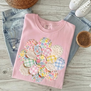 Peut inclure: Un sweat-shirt rose clair orné d'un grand motif floral composé de patchs de tissus variés. Les patchs présentent des motifs floraux, vichy et en dentelle. Le sweat-shirt est présenté avec un jean bleu clair, des baskets blanches et un petit panier tressé.