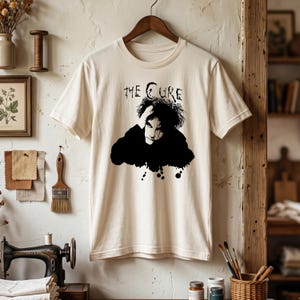 May include: A cream-colored short-sleeve t-shirt featuring a black graphic print of a person with messy hair and the text "THE CURE" above the figure. The shirt has a crew neckline and is displayed on a wooden hanger against a neutral wall.