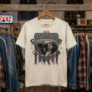 May include: A cream-colored short-sleeve t-shirt featuring a graphic print of the band Creedence Clearwater Revival. The design includes the text "CREEDENCE CLEARWATER REVIVAL" and "I AIN'T NO FORTUNATE SON" with a portrait of the band members.