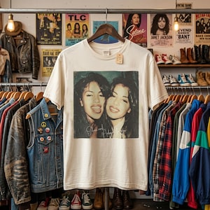 May include: A cream-colored short-sleeve t-shirt featuring a square graphic print of two women with the text "Selena and Aaliyah forever" below their faces. The shirt is displayed on a black hanger against a background of various clothing items.