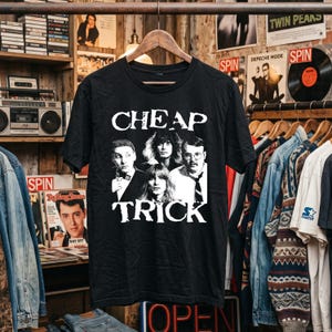 May include: A black short-sleeve t-shirt featuring a white graphic print of the band Cheap Trick. The design includes a black and white photograph of the four band members with the text CHEAP TRICK printed in a distressed white font above and below.