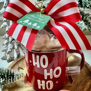 Puede incluir: Una taza roja esmaltada con la inscripción blanca "HO HO HO", llena de caramelos rosas, coronada con un lazo a rayas rojas y blancas. Una etiqueta verde con árboles de Navidad cuelga del lazo. Un bastón de caramelo está cerca.