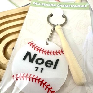 May include: Baseball-themed keychains with a white baseball design, red stitching, and a small wooden bat charm. Each keychain features a name and number, with the text "Noel 11" visible on one.
