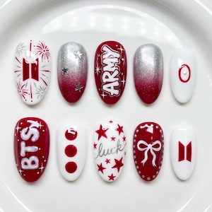 May include: A collection of ten artificial nails with diverse designs. The nails are in white, silver, and red. Some nails have the text "BTS" and "ARMY", while others display stars, bows, and other symbols. The nails are oval-shaped.