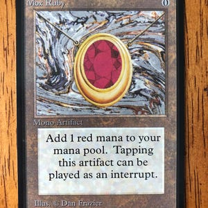 May include: A Mox Ruby card featuring a gold pendant with a large red gemstone. The card has the text "Add 1 red mana to your mana pool. Tapping this artifact can be played as an interrupt."