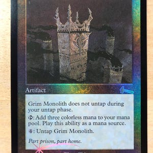 May include: A foil Magic: The Gathering trading card titled Grim Monolith. The card features a dark illustration of a stone tower structure, the text 'Artifact', game rules, the flavor text 'Part prison, part home.', and the illustrator name Chippy.