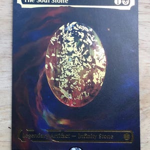 Op de afbeelding: Een rechthoekige verzamelkaart getiteld The Soul Stone met een centrale ovale oranje edelsteen met goudfolie accenten tegen een donkerblauwe en paarse kosmische achtergrond. De tekst bevat Legendary Artifact - Infinity Stone, M 0242 en Marvel.