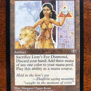 Może przedstawiać: Karta kolekcjonerska zatytułowana Lion's Eye Diamond z ilustracją osoby o ciemnych włosach w żółtej sukni, stojącej obok lwa i trzymającej biały klejnot wewnątrz czerwonego trójkąta. Tekst na karcie zawiera szczegółowe zasady gry.