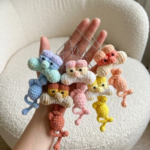 May include: A collection of five small, handmade crochet animal keychains, each featuring a distinct color combination including blue, pink, yellow, and peach. Each figure has a rounded head, small dark eyes, and a thin, dangling body with a metal chain.
