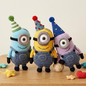 May include: Three crocheted Minion figures in blue, yellow, and purple, each wearing denim dungarees and a party hat. The Minions have large, round eyes and are posed on a wooden surface with small crocheted decorations.
