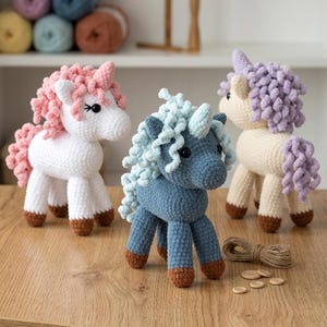 以下が含まれることがあります： ピンク、青、紫のたてがみを持つ、白、青、クリーム色の手作りの編み物ユニコーン3体。各ユニコーンは茶色の蹄と角を持っています。ユニコーンは木製の表面に展示されています。