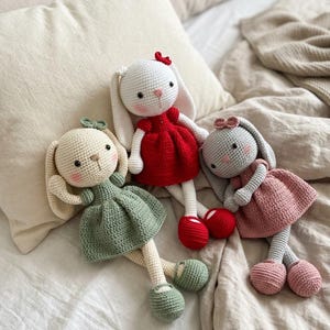 May include: Three handmade crochet bunny dolls. One wears a red dress and bow, another a green dress, and the third a pink dress. Each has matching shoes. The dolls are set on a bed with neutral-toned bedding.