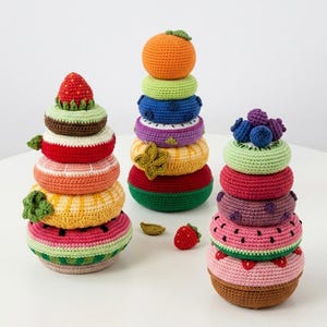 Può includere: Tre giocattoli impilabili all'uncinetto a forma di frutta. Uno è una fragola, uno un'arancia e uno un mirtillo. Ogni giocattolo è fatto di filato colorato e ha un design unico. I giocattoli sono su una superficie bianca.