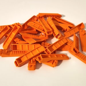May include: A pile of orange plastic shims, used for leveling or spacing. The shims are various shapes and sizes, with a bright, solid orange color. They are scattered across a white surface.