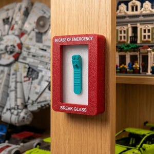 May include: A red rectangular emergency box with a glittery finish, featuring the white text IN CASE OF EMERGENCY and BREAK GLASS. Inside the box, mounted on a white foam backing, is a single teal plastic brick separator tool.