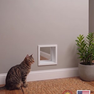 May include: A square white wall-mounted pet portal with a hollow center, featuring a clean frame design. The unit is installed on a neutral-colored wall, providing a clear opening for small animals to pass through, with a smooth finish.