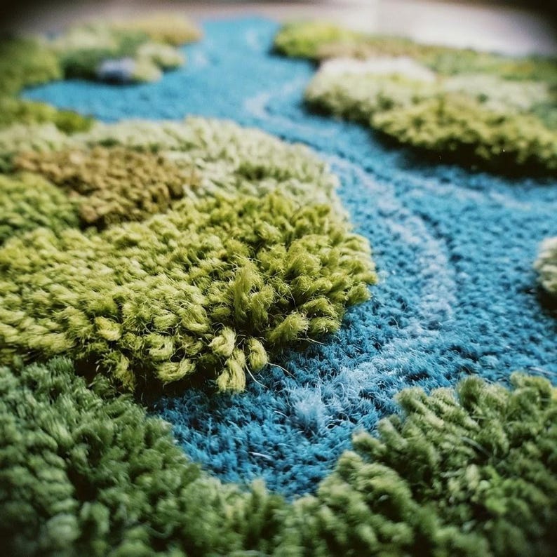May include: A close-up of a textured rug with a landscape design. The rug features a blue river winding through green and light green tufted areas, resembling islands or landmasses. The rug's texture is soft and plush.