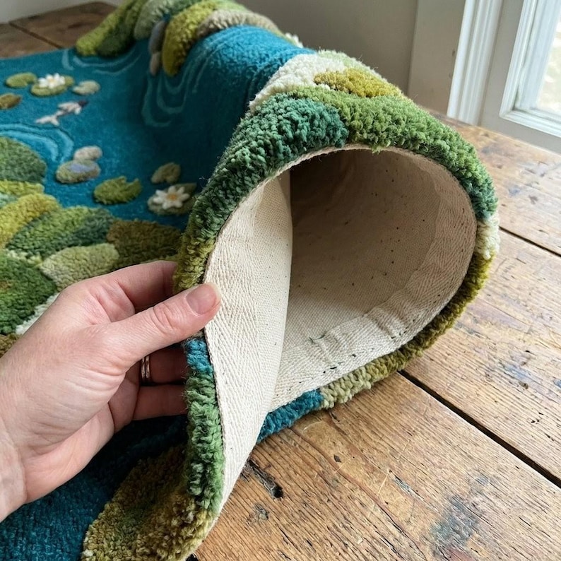 May include: A rolled-up rug with a pond scene featuring lily pads, flowers, and fish. The rug has a teal background with green and white accents. The back of the rug is a cream-colored fabric.