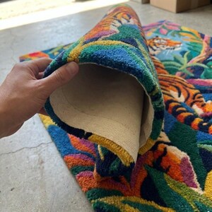 May include: A colorful rug with a tiger design. The rug features a vibrant jungle scene with orange, blue, green, and yellow hues. The rug is partially rolled up, revealing a beige backing.