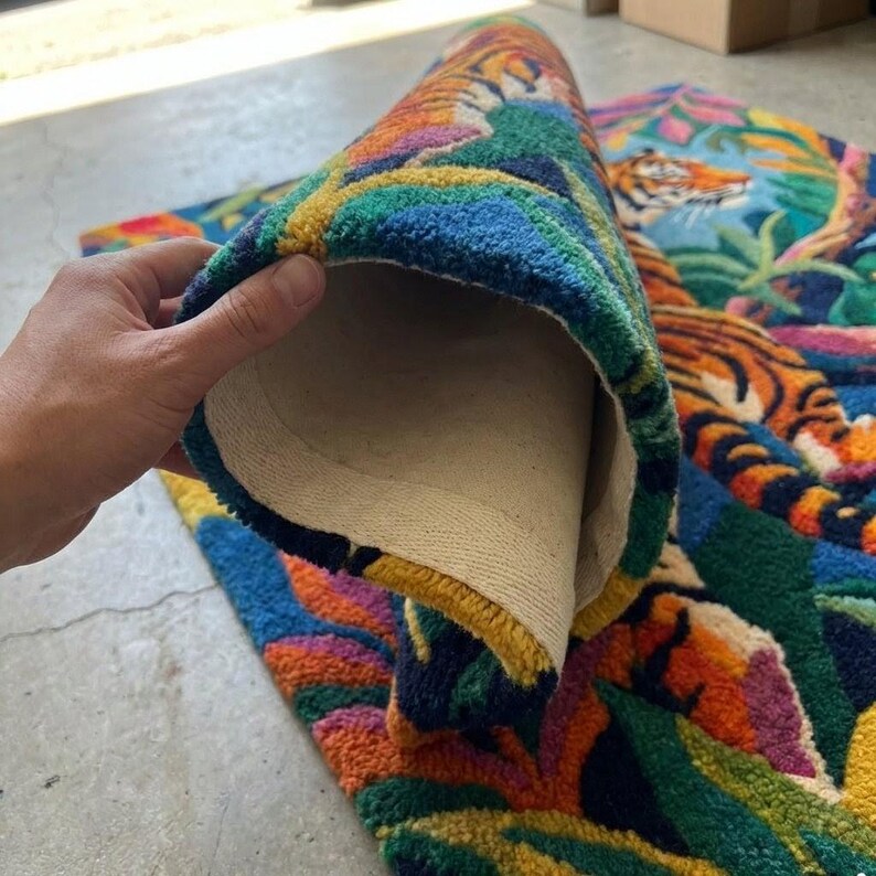 May include: A colorful rug with a tiger design. The rug features a vibrant jungle scene with orange, blue, green, and yellow hues. The rug is partially rolled up, revealing a beige backing.