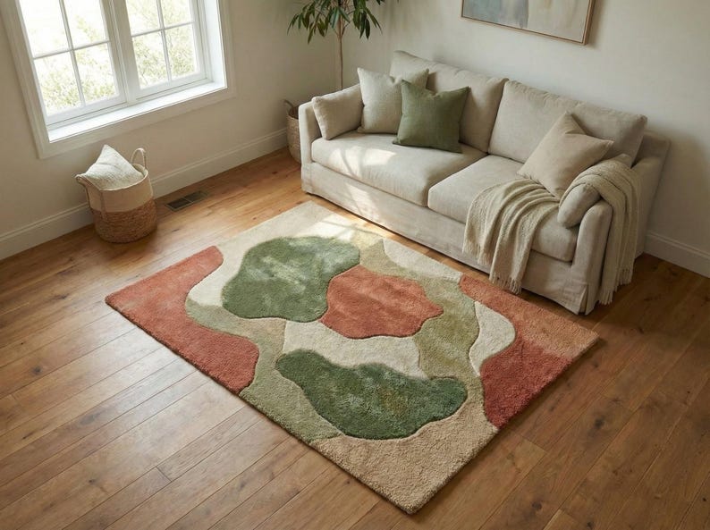 Puede incluir: Una alfombra rectangular con un dise&ntilde;o abstracto en tonos verdes, &oacute;xido y beige. La alfombra est&aacute; colocada sobre un suelo de madera frente a un sof&aacute; beige con cojines y una manta. Se ve una ventana y una cesta.