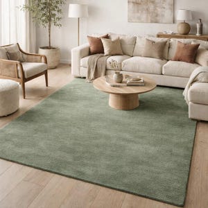 May include: A large, sage green area rug in a living room setting. The rug is rectangular and covers a significant portion of the hardwood floor. The room also features a white sofa, wooden coffee table, and a woven armchair.